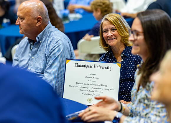 A woman holding an Emerita Status Certificate sitting in a crowd, posing for a photo